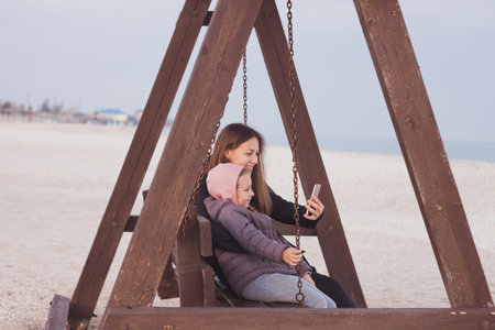 Happy mother and daughter sitting on wooden swing with smartphone autumn sea beach. Smiling...