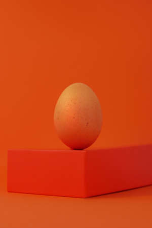 Easter egg on geometrical shape podium stage orange background. Brown chicken egg balancing on pedestal isometric minimalism. Creative minimal trendy monochrome colors still life. Modern composition.の写真素材