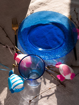 Easter table setting empty blue glass plate fork knife tree branches linen cloth top view harsh...