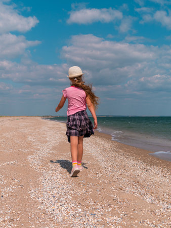 Happy child in a straw hat running jumping having fun on empty autumn sea beach. Blond girl walking on white sand road with blue cloudy sky background. Lifestyle photo Happiness freedom travel conceptの写真素材