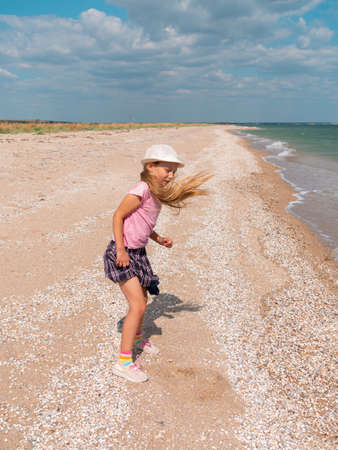 Happy child in a straw hat running jumping having fun on empty autumn sea beach. Blond girl walking on white sand road with blue cloudy sky background. Lifestyle photo Happiness freedom travel conceptの写真素材