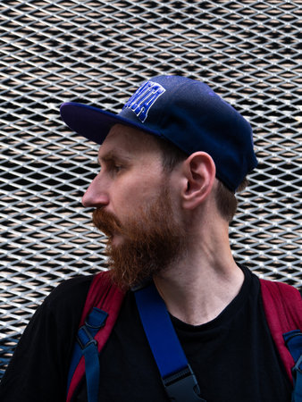 Profile portrait of bearded millennial hipster man in snapback on metal fence urban background. Stylish confident authentic guy with red beard casual clothes backpack walking on the sunny city street.の写真素材