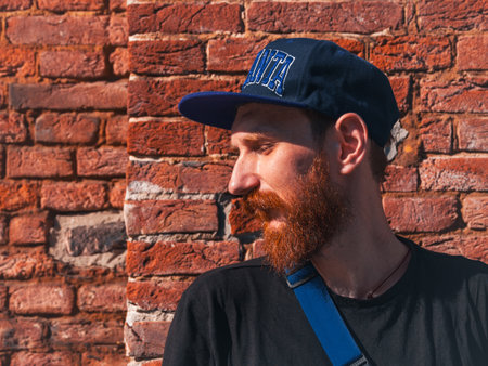 Profile portrait of bearded millennial hipster man in snapback on brick wall urban background. Stylish confident authentic guy with red beard casual clothes black t-shirt walking on sunny city street.の写真素材