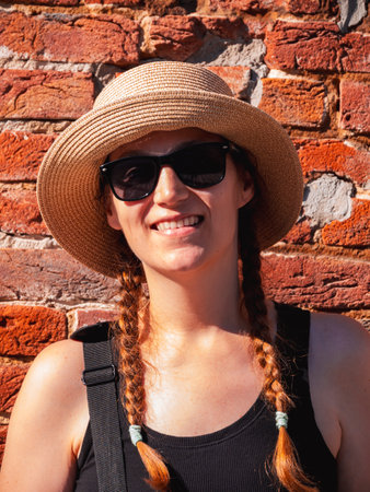 Young woman in straw hat portrait on red brick wall urban background. Millennial girl walking on summer city street with bright sunshine. Stylish confident authentic lady in black sunglasses outdoors.の写真素材