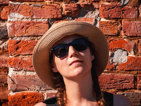 Young woman in straw hat portrait on red brick wall urban background. Millennial girl walking on summer city street with bright sunshine. Stylish confident authentic lady in black sunglasses outdoors.の写真素材