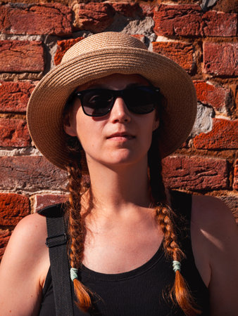 Young woman in straw hat portrait on red brick wall urban background. Millennial girl walking on summer city street with bright sunshine. Stylish confident authentic lady in black sunglasses outdoors.の写真素材