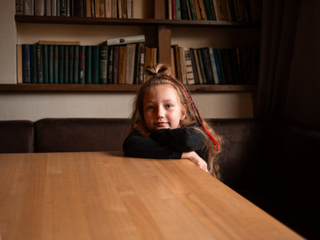 Adorable stylish little girl with long colorful hair in modern library cafe book shelf background. Young reader sitting alone on comfortable couch at the table Authentic cheerful candid child portraitの写真素材