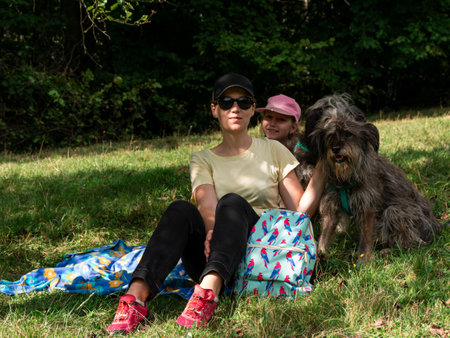 Happy family mother with daughter and two dogs sitting on green grass lawn having fun picnic spending time together hiking in the mountains. People and pets outdoors lifestyle photo candid emotions.の写真素材