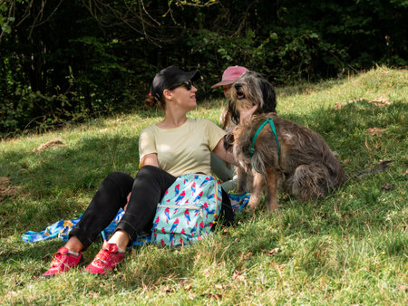Happy family mother with daughter and two dogs sitting on green grass lawn having fun picnic spending time together hiking in the mountains. People and pets outdoors lifestyle photo candid emotions.の写真素材