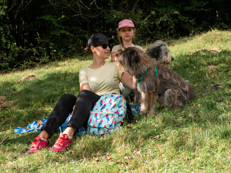 Happy family mother with daughter and two dogs sitting on green grass lawn having fun picnic spending time together hiking in the mountains. People and pets outdoors lifestyle photo candid emotions.の写真素材