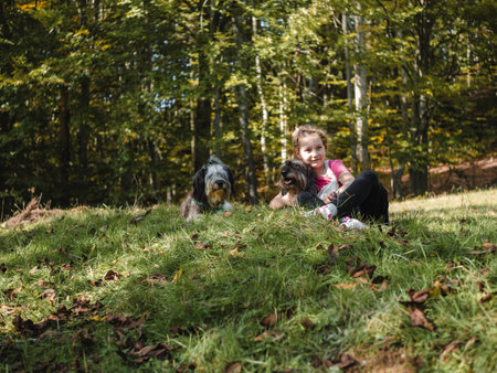 Happy little girl with two dogs sitting on a green grass lawn having fun, family picnics, spending time together, hiking in the mountains. People and pets outdoors lifestyle photo candid emotions.の写真素材