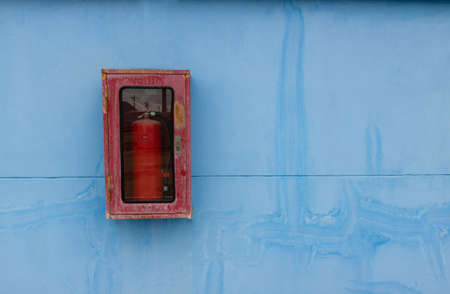 The old red fire extinguisher is in the old cabinet, rusting Installed on the blue warehouse wall.の写真素材
