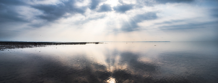 Sun breaking through the clouds as Whitstable beach is at low tide creating a calm reflection of the skyの写真素材