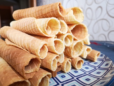 Close-up of delicious chocolate waffle rolls lie on white plate. Preparation of a sweet dessert. White clean kitchen and table. Many wafer tubules are group. Edible tubes with white spiral from cream. Waffle pipes on a blue plateの写真素材