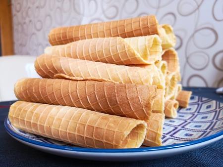 Close-up of delicious chocolate waffle rolls lie on white plate. Preparation of a sweet dessert. White clean kitchen and table. Many wafer tubules are group. Edible tubes with white spiral from cream. Waffle pipes on a blue plateの写真素材