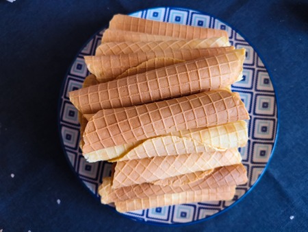 Close-up of delicious chocolate waffle rolls lie on white plate. Preparation of a sweet dessert. White clean kitchen and table. Many wafer tubules are group. Edible tubes with white spiral from cream. Waffle pipes on a blue plateの写真素材