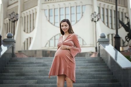 Pregnant girl posing in the city. young pregnant woman posing on the street at the theater, stroking her stomach. Enjoying the state of pregnancy and motherhood woman in a beige light dress.の写真素材