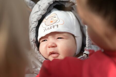 A girl in warm clothes on the street. A young and happy mother holds a baby in her arms, smiles and rejoices. Street in winter, fresh air, a boy up to a year. Winter warm clothes, weekend holiday concept at winter resort. jumpsuit with ears.の写真素材