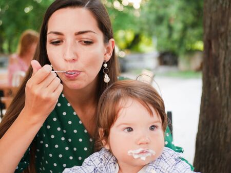mom feeds her baby on the street. A mother feeds her little baby with a spoon on the street. mouth in food. healthy cottage cheese and yogurd.の写真素材