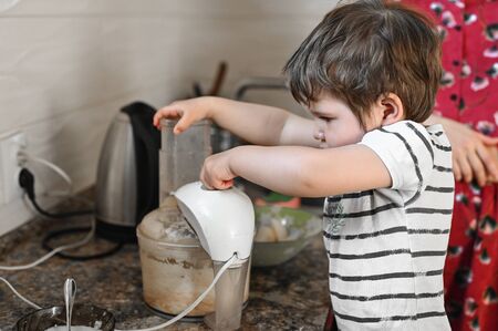Little child helps mom in the kitchen with cooking, whipping food in a combine.の写真素材