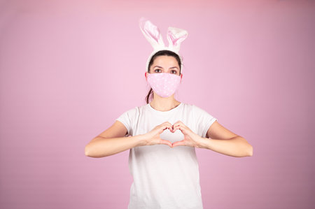 Young age woman wearing coronavirus protection mask doing heart symbol shape with hands on pink background. Easter concept. High quality photoの写真素材