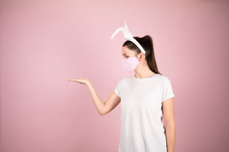 Young woman wearing bunny ears and protective mask standing over isolated pink background cheerful presenting and pointing with palm at something. High quality photoの写真素材