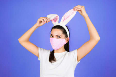 A young woman in the ears of an Easter Bunny, wearing a medical protective mask on her face isolated...