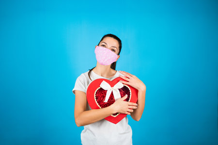 Woman in pink protective mask hold large heart shaped gift box. Holidays presents, shopping sale during the pandemic. Blue background with copy space. High quality photoの写真素材