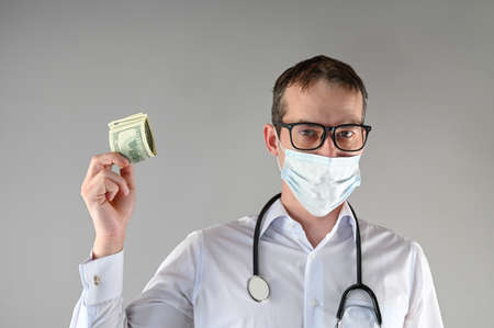 A male doctor in a mask holds money in his hand, on an isolated background. High quality photoの写真素材