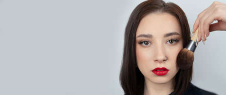 Young woman with makeup brush near at face. Beautiful girl with perfect skin, red lips and a pierced noseの写真素材