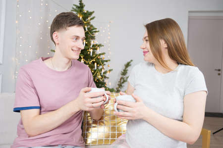 Loving couple celebrates Christmas. Pregnant woman holding cup with hot drink. Christmas tree in background. Happy holidaysの写真素材