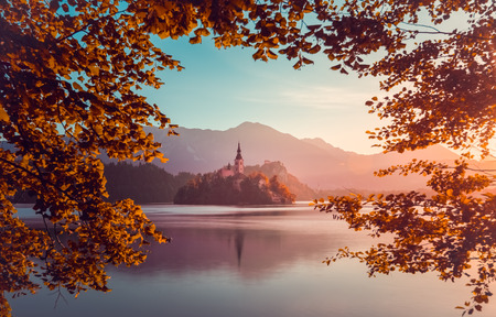 Little Island with Catholic Church in Bled Lake, Slovenia  at Sunrise with Castle and Mountains in Background. Autumn Filter. Tree Leaves Border. Natural Frame.の写真素材
