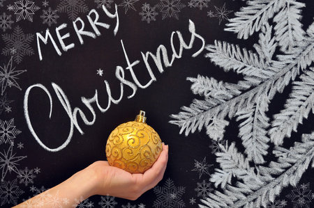 the female hand holds a Christmas ball on a chalkboard with an inscription Merry Christmas and fir-tree branches and snowflakesの写真素材