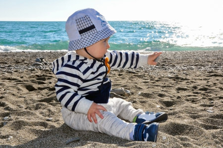 the one-year-old kid in a hat sitting on the beach also holding a pebble in handの写真素材