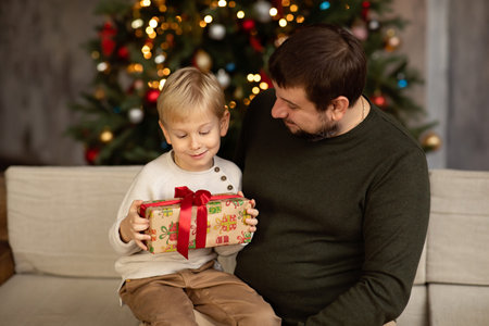 happy father and son with christmas gift box on sofa at home, christmas tree in backgroundの写真素材