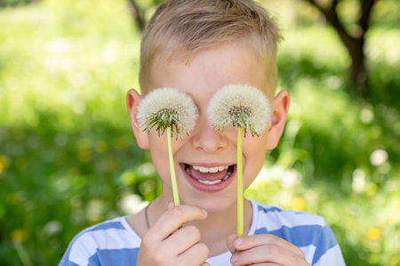 The boy has covered his eyes with dandelions and is smiling widely against a green background. Selective focus.の写真素材