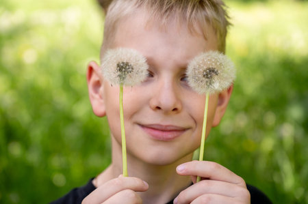 The boy has covered his eyes with dandelions and is smiling widely against a green background. Selective focus.の写真素材
