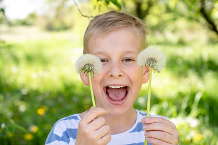 The boy has covered his eyes with dandelions and is smiling widely against a green background. Selective focus.の写真素材