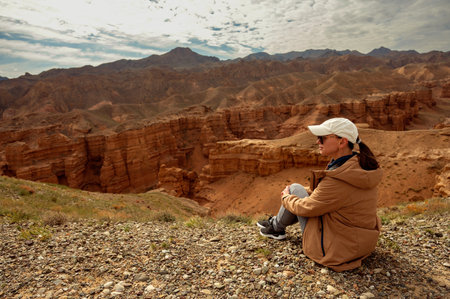 female tourist sits on a hill and looks at the beautiful orange canyon mountains and cloudy skyの写真素材