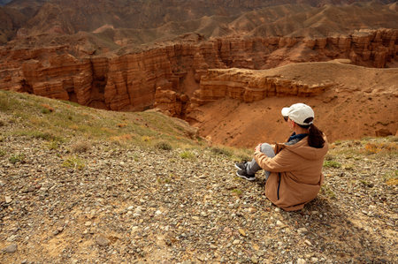 female tourist sits on a hill and looks at the beautiful orange canyon mountains and cloudy skyの写真素材