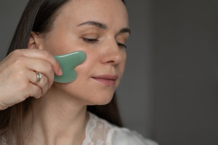 Young woman using a green gua sha scraper for facial massage, improving skin circulation and relaxation. Home skincare and self-care routine for beauty and wellness.の写真素材