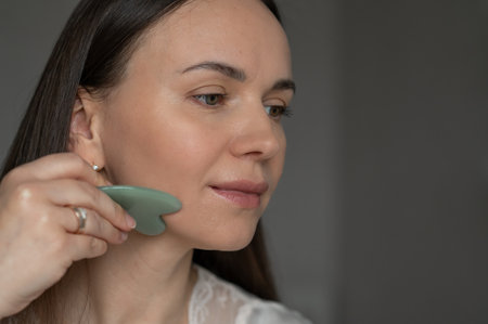 Young woman using a green gua sha scraper for facial massage, improving skin circulation and relaxation. Home skincare and self-care routine for beauty and wellness.の写真素材