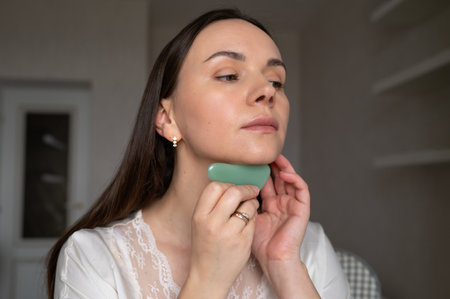 Young woman in silk robe doing facial massage with gua sha scraper while looking in mirror. Home skin care and aesthetic proceduresの写真素材