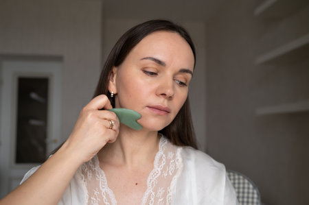 Young woman in silk robe doing facial massage with gua sha scraper while looking in mirror. Home skin care and aesthetic proceduresの写真素材