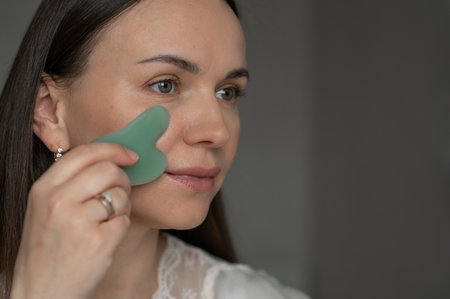 Young woman using a green gua sha scraper for facial massage, improving skin circulation and relaxation. Home skincare and self-care routine for beauty and wellness.の写真素材