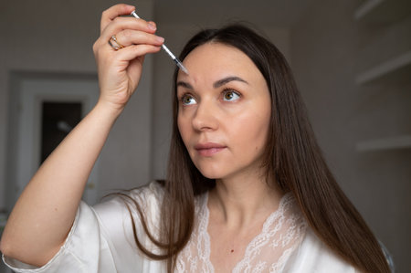 Young woman in robe giving beauty injection to her face looking in mirror. Home skin care and aesthetic proceduresの写真素材