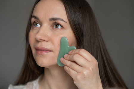Young woman using a green gua sha scraper for facial massage, improving skin circulation and relaxation. Home skincare and self-care routine for beauty and wellness.の写真素材