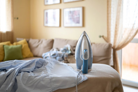 Steam iron on ironing board and pile of crumpled laundry in background in bright cozy living room.の写真素材