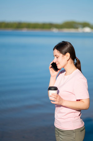 Young woman drinking coffee and talking on a smartphone by the lake shore on a sunny day.の写真素材