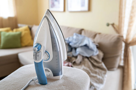Electric steam iron on ironing board and pile of wrinkled laundry on sofa in background in cozy bright living rooの写真素材